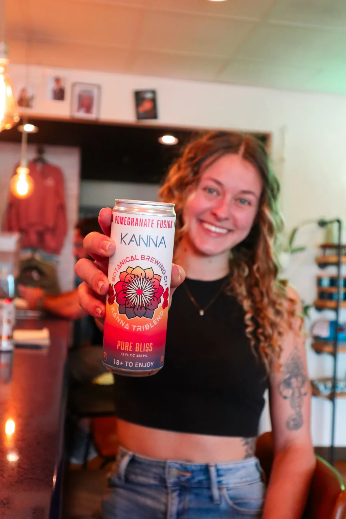 A smiling woman holding up a can of Kanna Pomegranate Fusion at a bar